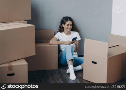 Happy latina female uses mobile phone apps, choosing moving service, preparing for relocation, answer her mover online, sitting on the floor with packed things in cardboard boxes. Easy moving day. Happy girl uses phone apps, choosing relocation service, answers her mover online. Easy moving day