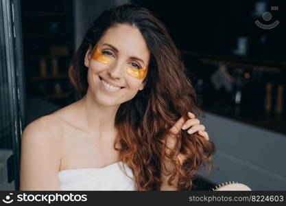 Happy latina woman applies eye patches for reducing wrinkles brushing healthy curly long hair at home. Smiling female combing hairs after shower. Morning beauty routine. Skincare, haircare, self-care.. Latina girl with under eye patches brushing hair after shower. Skincare, haircare, self care routine