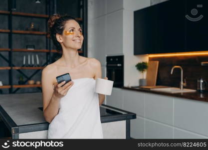Happy latina woman wrapped in towel with hydrogel under eyes patches holding smartphone and morning coffee at home. Smiling dreaming young female after shower got good news message in social networks.. Happy woman with under eye patches holding smartphone and morning coffee got good news message