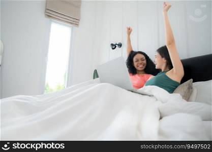 Happy lesbian couple using laptop on bed at home. Young multiethnic lesbian couple using laptop together in bedroom. Technology and love concept