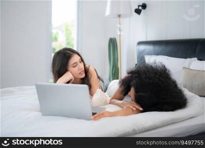 Happy lesbian couple using laptop on bed at home. Young multiethnic lesbian couple using laptop together in bedroom. Technology and love concept