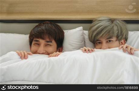 Happy LGBT Asian young gay couple with eyes smiling popped half of his face out of the blanket in bedroom at home together