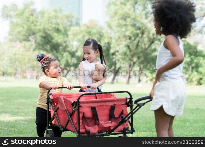 Happy little African American and Caucasian kids girls playing together at park in summer. Children towing and pushing red trolley car on grass to camping. Friendship of diverse ethnicity children