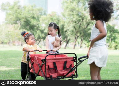 Happy little African American and Caucasian kids girls playing together at park in summer. Children towing and pushing red trolley car on grass to camping. Friendship of diverse ethnicity children