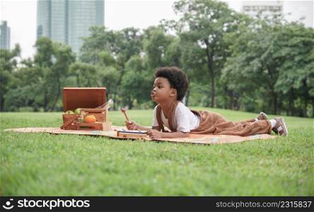 Happy little African boy lying down on mat and playing xylophone toy at garden. African family picnic together at park with basket of fruits and drinks in glass bottle on sunny day