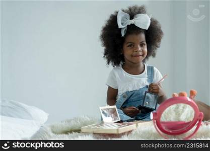 Happy little African girl playing makeup with mother?s cosmetics. Adorable kid holding colors palette and pink brush painting her face with joy in bedroom at home