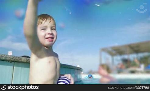 Happy little boy having fun in swimming pool on resort. He splashing water on the camera and laughing