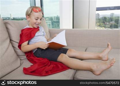 Happy little boy in superhero costume reading book on sofa at home
