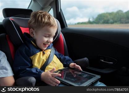 Happy little boy in the car sitting in child safety seat and playing with tablet PC