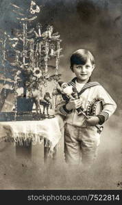 Happy little boy with christmas tree, gifts and vintage toys. Antique sepia picture with original film grain and scratches