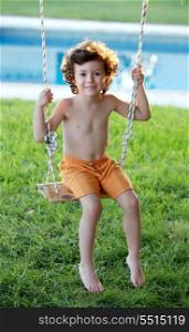 Happy little boy with curly hair sitting on a swing with a pool background