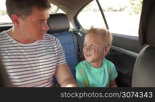 Happy little boy with toy and backpack traveling with young father on the backseat of a car. They having cheerful talk during ride