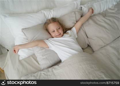 Happy little elementary child girl stretching, lying in soft comfortable bed after wake up. Smiling caucasian preschooler kid enjoying morning in cozy bedroom. Healthy children&rsquo;s sleep concept.. Happy little child girl stretching, lying in soft bed after wake up. Healthy children&rsquo;s sleep