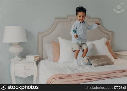 Happy little energetic afro american kid boy jumping alone on his parents bed while holding delicious lollipop, eating sweets, causing mess while no one is home, cute child having fun at home. Energetic afro american kid jumping with lollipop on bed mattress
