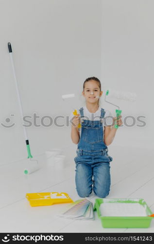 Happy little girl in denim overalls stands on knees, holds paint roller, surrounded with trays with paint, poses in white room, has happy expression, helps parents with house improvement and painting