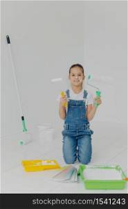 Happy little girl in denim overalls stands on knees, holds paint roller, surrounded with trays with paint, poses in white room, has happy expression, helps parents with house improvement and painting