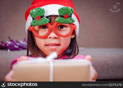 Happy little girl in Santa hat giving a Christmas present at home. Happy New Year and Merry Christmas.