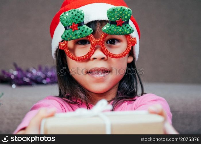 Happy little girl in Santa hat giving a Christmas present at home. Happy New Year and Merry Christmas.