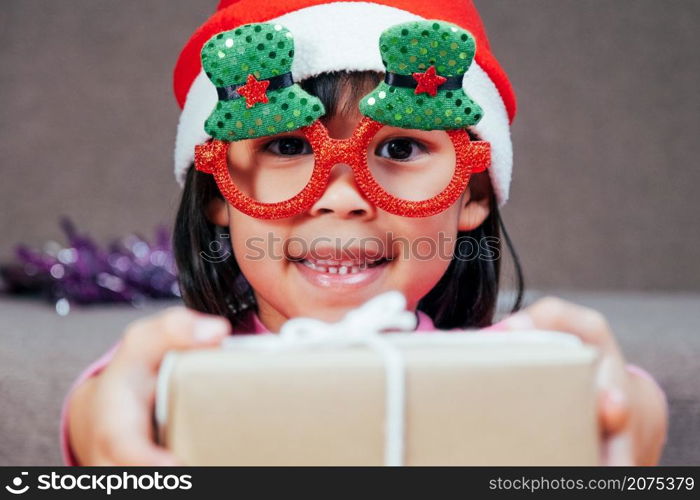 Happy little girl in Santa hat giving a Christmas present at home. Happy New Year and Merry Christmas.