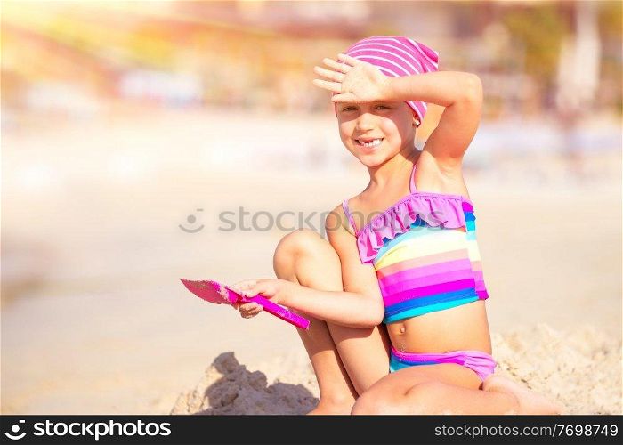 Happy little girl playing on the beach with shovel and sandbag in bright sunny day, active childhood, having fun outdoors, enjoying summer holidays on the seashore