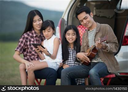 Happy little girl playing ukulele with asian family sitting in the car for enjoying road trip and summer vacation in camper van