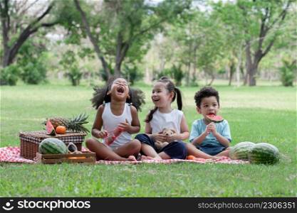 Happy little kids African, Caucasian and Asian picnic in the park. They laughing and sitting on grass eating watermelon with basket of fruits. Diverse ethnicity children friends enjoy life on summer