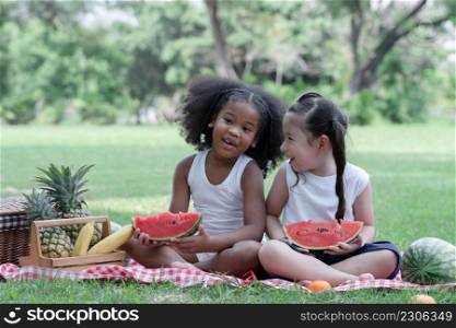 Happy little kids girl African and Caucasian eating watermelon while talking and sitting on grass picnic in the park with basket of fruits. Diverse ethnicity children friends enjoy life on summer