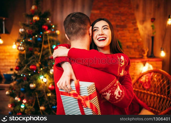 Happy love couple hugs, christmas holidays. Xmas celebration, cheerful man and woman with gift, fir-tree with decoration on background. Happy love couple hugs, christmas holidays