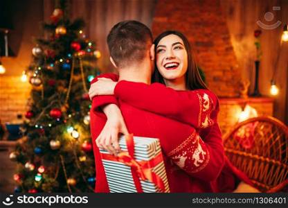 Happy love couple hugs, christmas holidays. Xmas celebration, cheerful man and woman with gift, fir-tree with decoration on background. Happy love couple hugs, christmas holidays