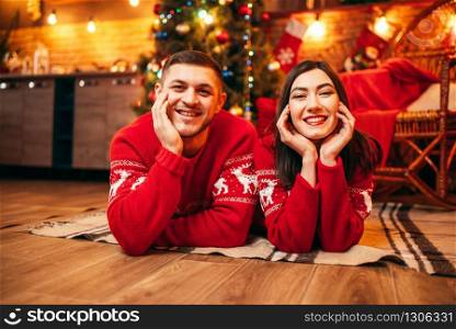 Happy love couple lies on the floor, christmas holidays. Xmas celebration, cheerful man and woman in red pullovers, fir-tree with decoration on background. Love couple lies on the floor, christmas holidays