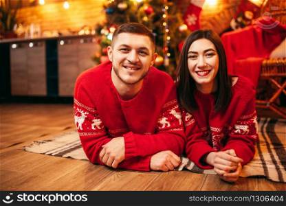 Happy love couple lies on the floor, christmas holidays. Xmas celebration, cheerful man and woman in red pullovers, fir-tree with decoration on background