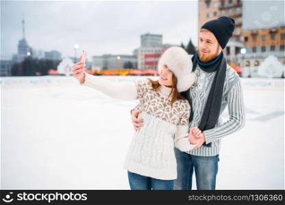Happy love couple makes selfie on skating rink. Winter ice-skating on open air, active leisure, man and woman skates together. Happy love couple makes selfie on skating rink