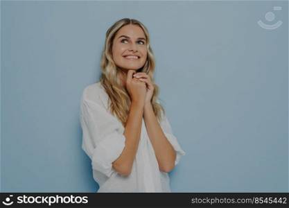 Happy lovely blonde girl feeling nostalgic recalling romantic memories, holding hands together and looking up with smile while standing isolated on blue studio background with copy space. Happy lovely blonde girl feeling nostalgic and looking up with smile