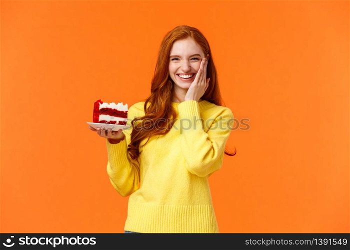 Happy lovely caucasian redhead female holding tasty cake with cream, touch cheek as receive congratulations being birthday girl, celebrating b-day with family, standing orange background.. Happy lovely caucasian redhead female holding tasty cake with cream, touch cheek as receive congratulations being birthday girl, celebrating b-day with family, standing orange background