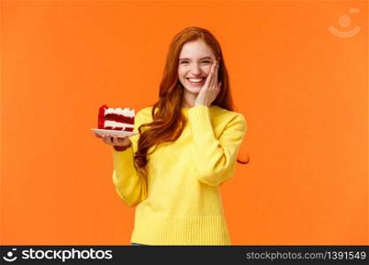 Happy lovely caucasian redhead female holding tasty cake with cream, touch cheek as receive congratulations being birthday girl, celebrating b-day with family, standing orange background.. Happy lovely caucasian redhead female holding tasty cake with cream, touch cheek as receive congratulations being birthday girl, celebrating b-day with family, standing orange background