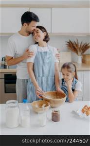 Happy lovely family in home kitchen, father embraces mother with love, little girl looks in bowl, observes how mommy cooks and whisks ingredients, use eggs for making dough. Domestic atmosphere