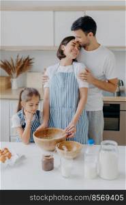 Happy lovely family in home kitchen, father embraces mother with love, little girl looks in bowl, observes how mommy cooks and whisks ingredients, use eggs for making dough. Domestic atmosphere