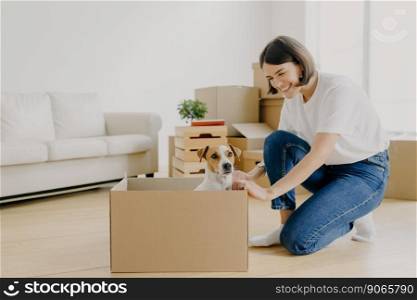 Happy lovely woman dressed in casual clothes, plays with favourite pet who poses in carton box, pose in white spacious room, lives in rented house express positivity. Moving Day and relocation concept