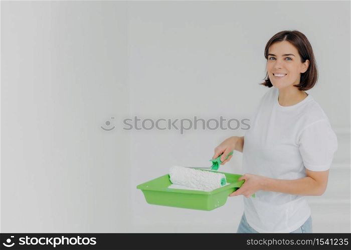 Happy lovely woman in white casual t shirt, poses with paint roller and special tray, makes refurbishment on new apartment, has short dark hair and toothy smile, isolated over white background.