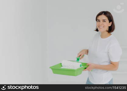 Happy lovely woman in white casual t shirt, poses with paint roller and special tray, makes refurbishment on new apartment, has short dark hair and toothy smile, isolated over white background.