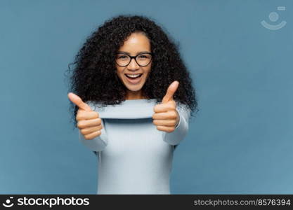 Happy lovely woman with bushy Afro hair gives thumb up, approves nice idea, wears fashionable sweater, isolated over blue background. Its fine, I agree. Body language concept. People and like concept