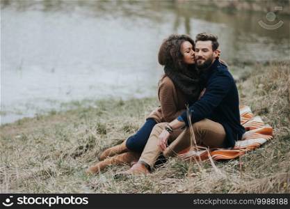 Happy lovers are sitting on the shore of the lake. Young couple is hugging on autumn day outdoors. A bearded man and curly woman in love. Valentine’s Day. Concept of love and family. Happy lovers are sitting on the shore of the lake. Young couple is hugging on autumn day outdoors. A bearded man and curly woman in love. Valentine’s Day. Concept of love and family.