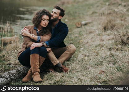 Happy lovers are sitting on the shore of the lake. Young couple is hugging on autumn day outdoors. A bearded man and curly woman in love. Valentine’s Day. Concept of love and family. Happy lovers are sitting on the shore of the lake. Young couple is hugging on autumn day outdoors. A bearded man and curly woman in love. Valentine’s Day. Concept of love and family.