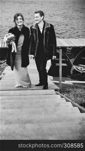 Happy loving couple holding hands standing on the bridge against the blurred lake with boats. Selective focus, black and white vintage film filter.. Happy Couple In Monochrome