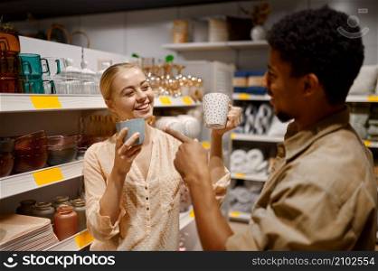Happy loving just married couple choosing ceramic cups at household goods supermarket department. Loving couple choosing ceramic cups at shop