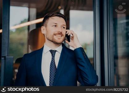 Happy male business owner in blue suit receiving good news about project results via smartphone, smiling and listening with pleasure while standing next to glass entrance door of office building. Male business owner hearing good news over phone