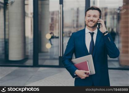 Happy male company worker leaving office building holding notebook and laptop while talking on phone with his colleague, sharing good news about recent events, blurred background. Smiling male company worker leaving office building while talking on phone with his colleague
