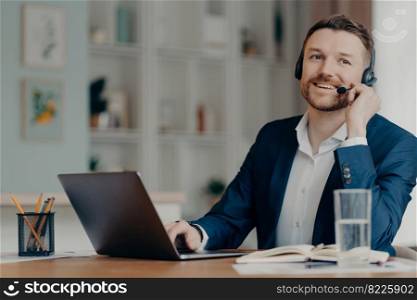 Happy male entrepreneur in suit working from home, successful businessman in headset talking with colleagues online through web conference while sitting at his cozy workplace. Remote work at home. Smiling manager having online call on laptop while sitting at table in living room