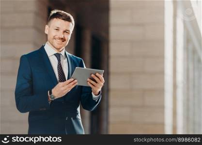 Happy male entrepreneur wears formal black suit, holds modern tablet computer, checks financial news in networks, connected to wireless internet, prepares financial report on business meeting