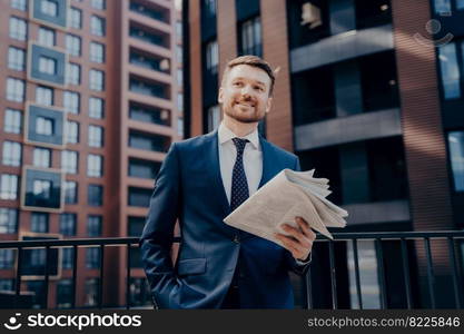 Happy male executive in blue suit with newspaper reading latest news, trying to find new ways to make profit, standing alone with hand in his pocket and plaza in background. Successful people. Happy ceo in blue suit with newspaper next to office buildings